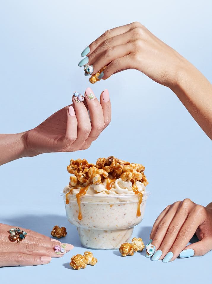 Hands scattering caramel popcorn over a cup of Four Winters ice cream against a pale blue background